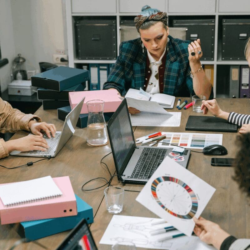 Four professionals collaborate at a wooden table during a creative brainstorming session, reviewing design samples and color palettes for glass decorating and product customization projects