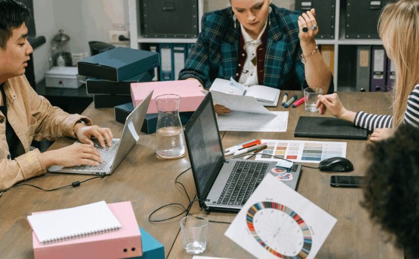 Four professionals collaborate at a wooden table during a creative brainstorming session, reviewing design samples and color palettes for glass decorating and product customization projects