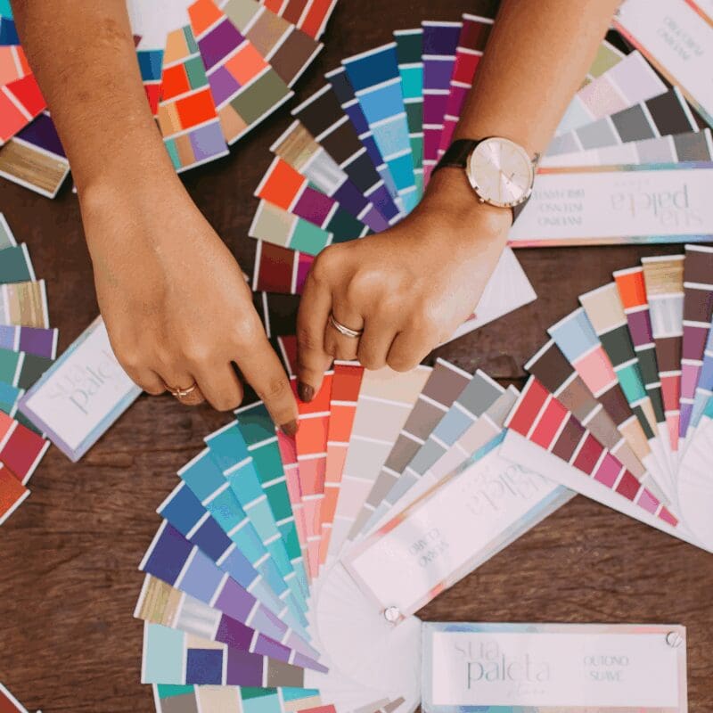 Person selecting paint colors from multiple fan-style color swatches spread across a wooden table with paint samples and design materials