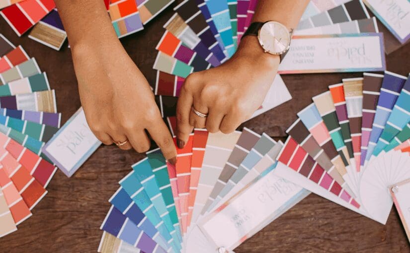 Person selecting paint colors from multiple fan-style color swatches spread across a wooden table with paint samples and design materials