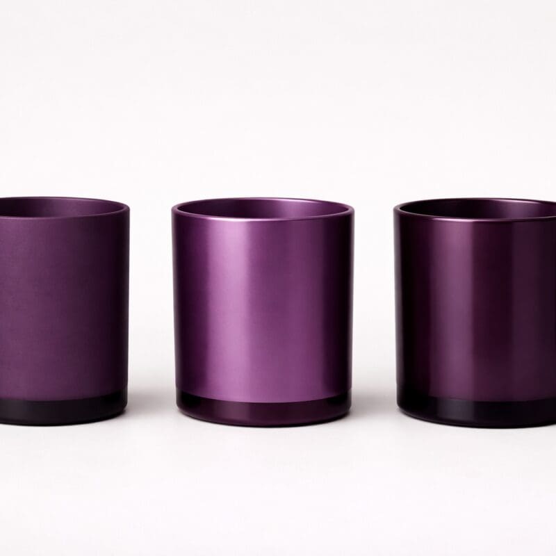 Three purple metallic cylindrical candle jars with black bases arranged in a row against a white background