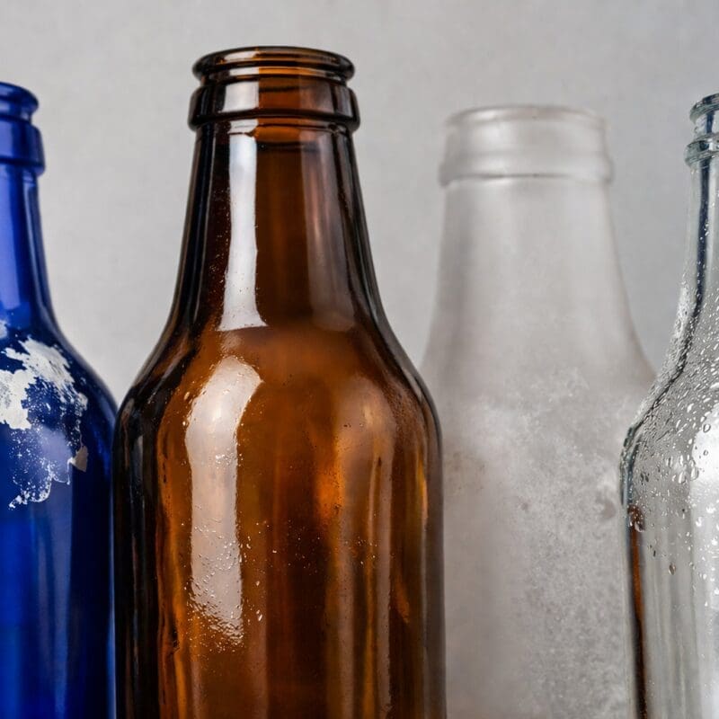 Four glass bottles in blue, brown, frosted, and clear with varying surface finishes and condensation, showcasing glass decorating and customization options