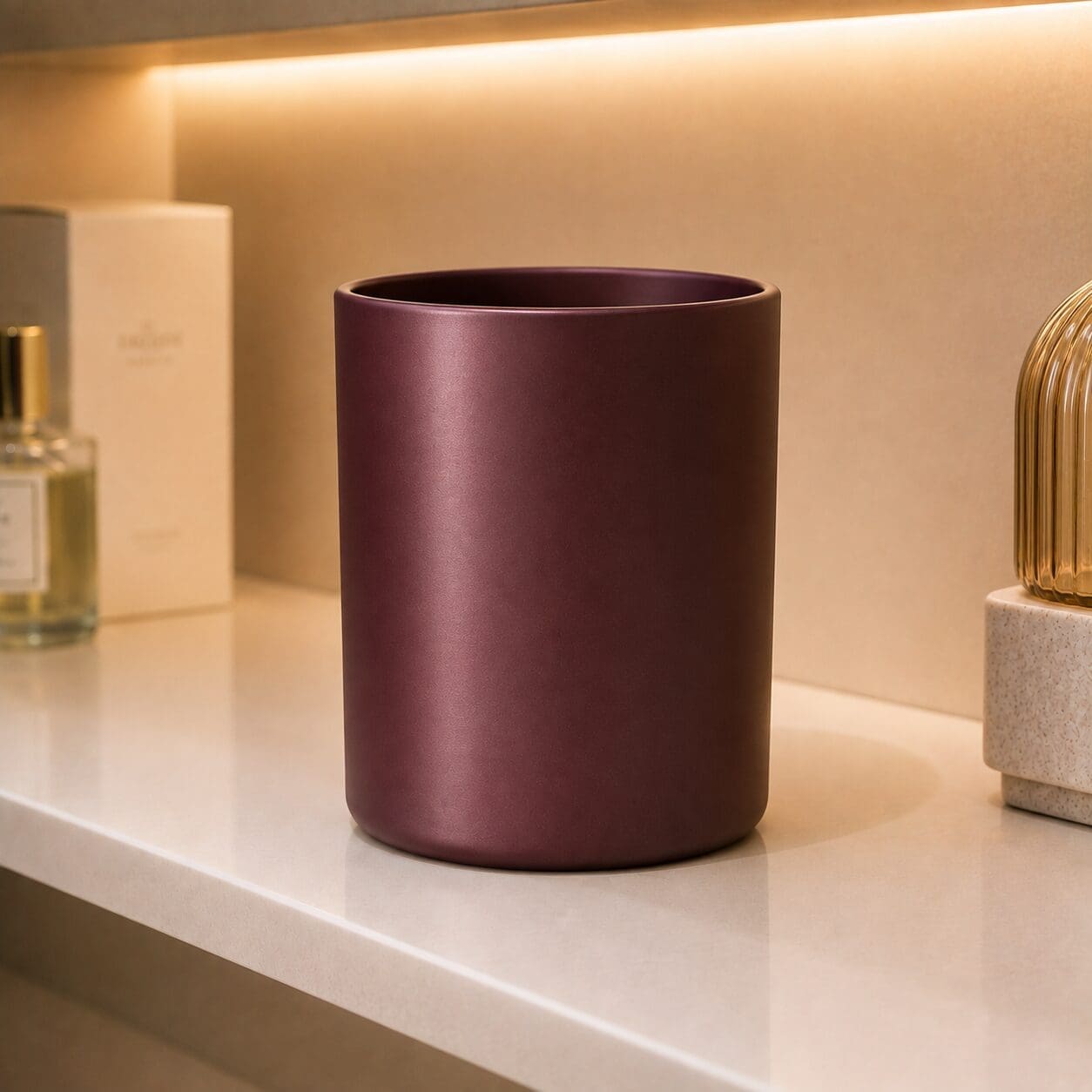 Burgundy matte cylindrical candle jar displayed on white shelf among perfume bottles and decorative items with warm ambient lighting