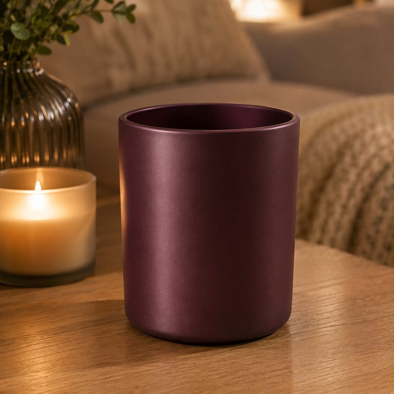 Empty burgundy matte cylindrical glass jar on wooden table surrounded by lit candle, greenery in ribbed gold vase, and blurred home decor