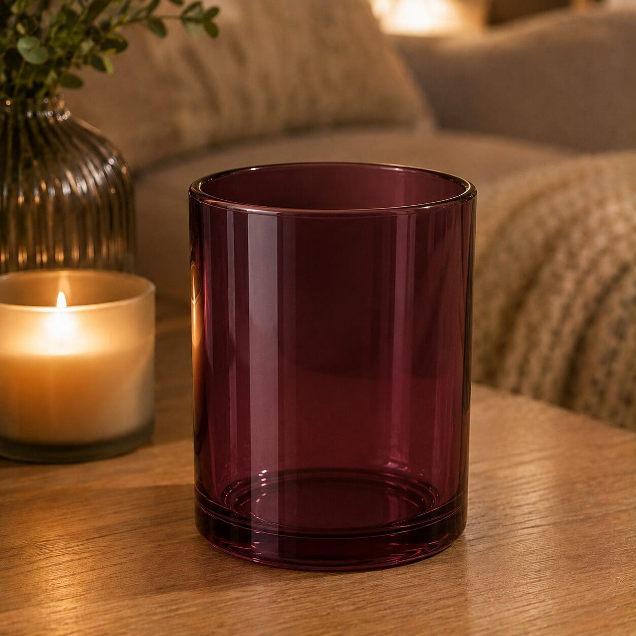 Deep burgundy glass cylinder vase on wooden table with lit candle and greenery in background, showcasing decorative glassware styling