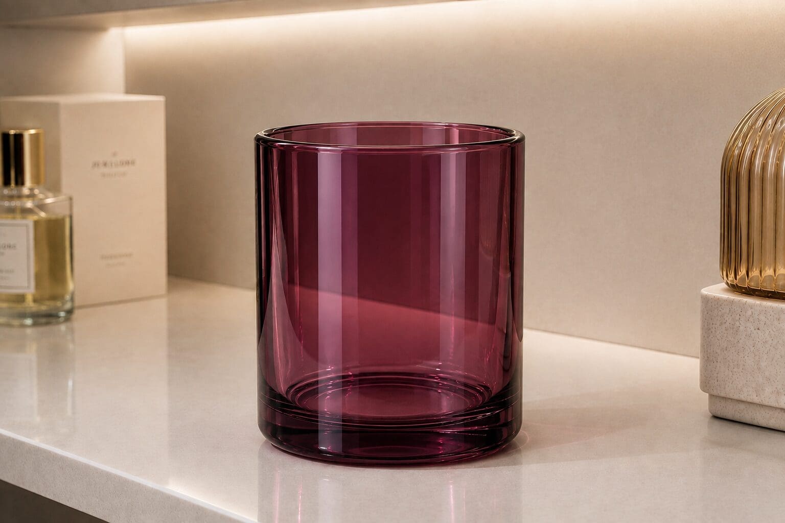Deep burgundy glass candle jar displayed on white shelf with perfume bottle and decorative gold-ribbed vase nearby