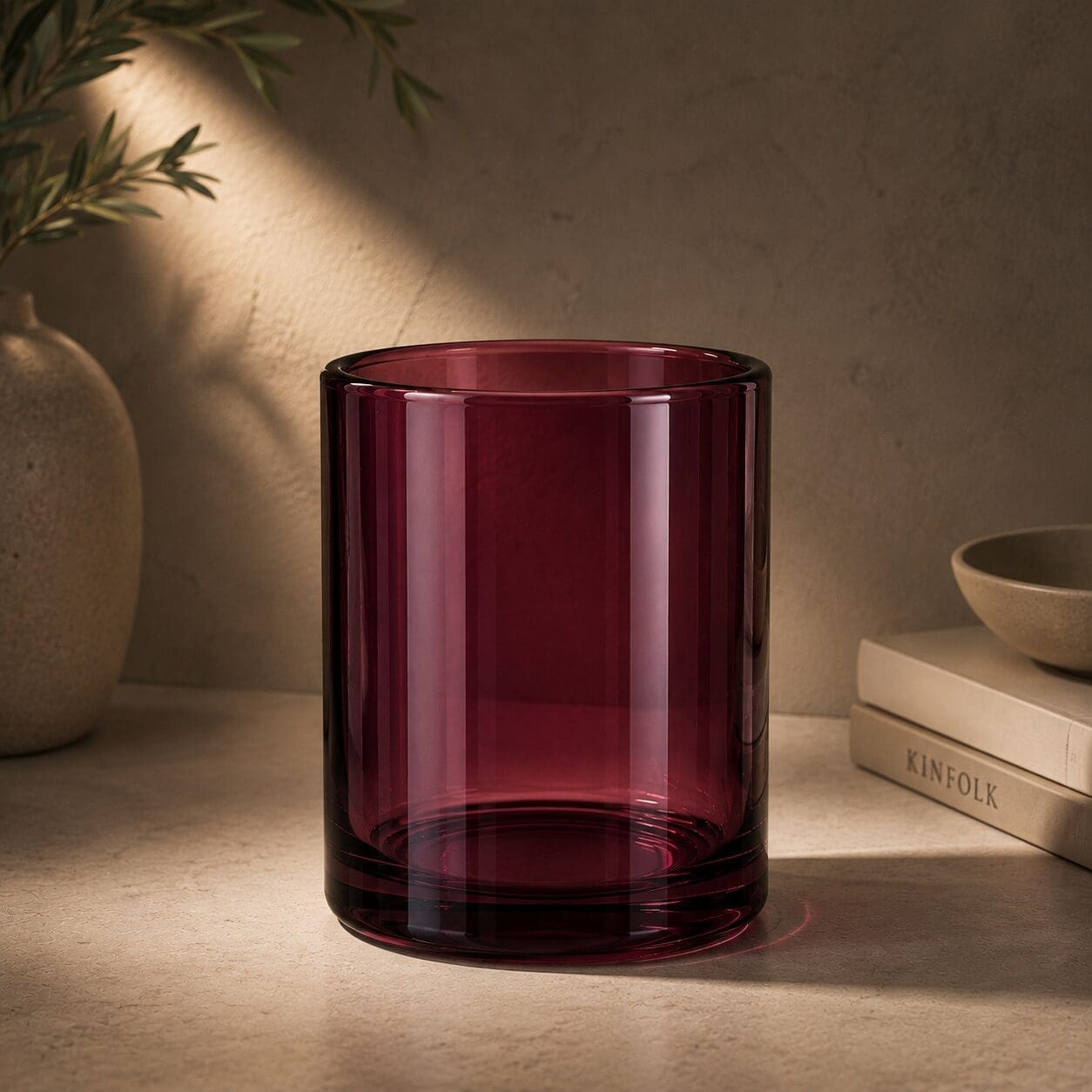 Deep burgundy glass cylinder vase on beige surface with natural sunlight, surrounded by ceramic bowl, plant, and "Kinfolk" book