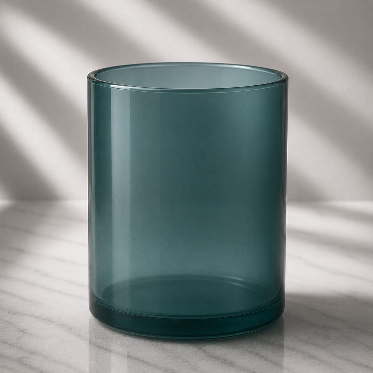 Empty teal glass cylinder on marble surface with sunlight casting shadows in background