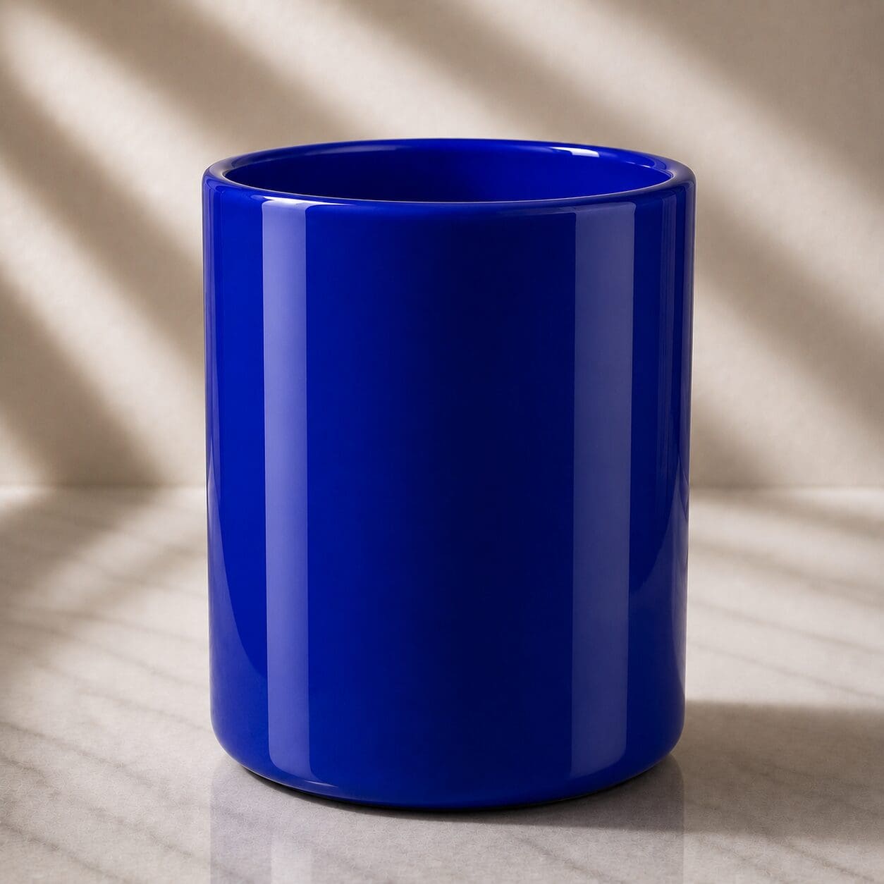Bright cobalt blue ceramic mug with glossy finish, displayed on marble surface with natural light creating shadows in background