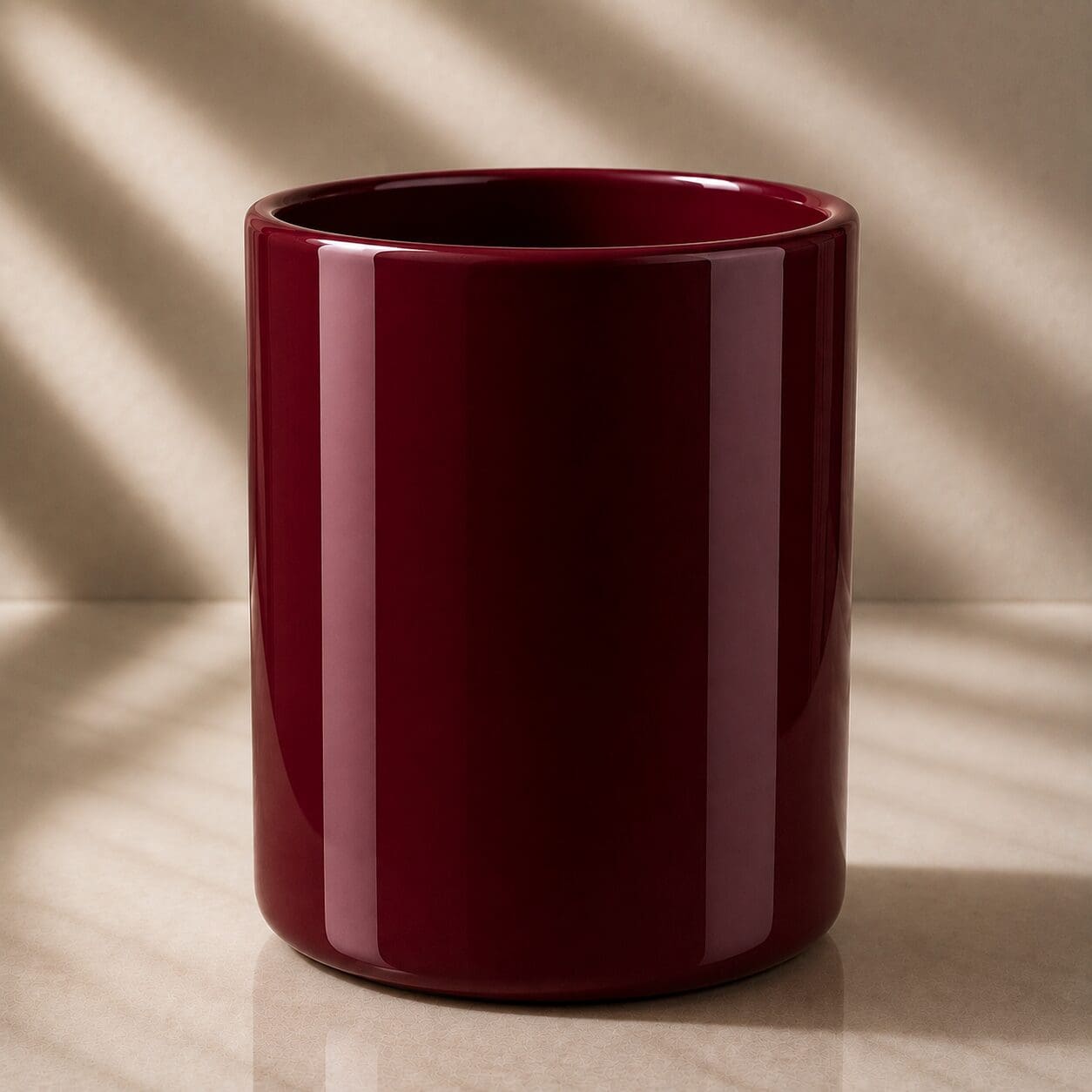 Deep burgundy cylindrical glass jar with glossy finish, shown on neutral beige surface with natural light casting diagonal shadows