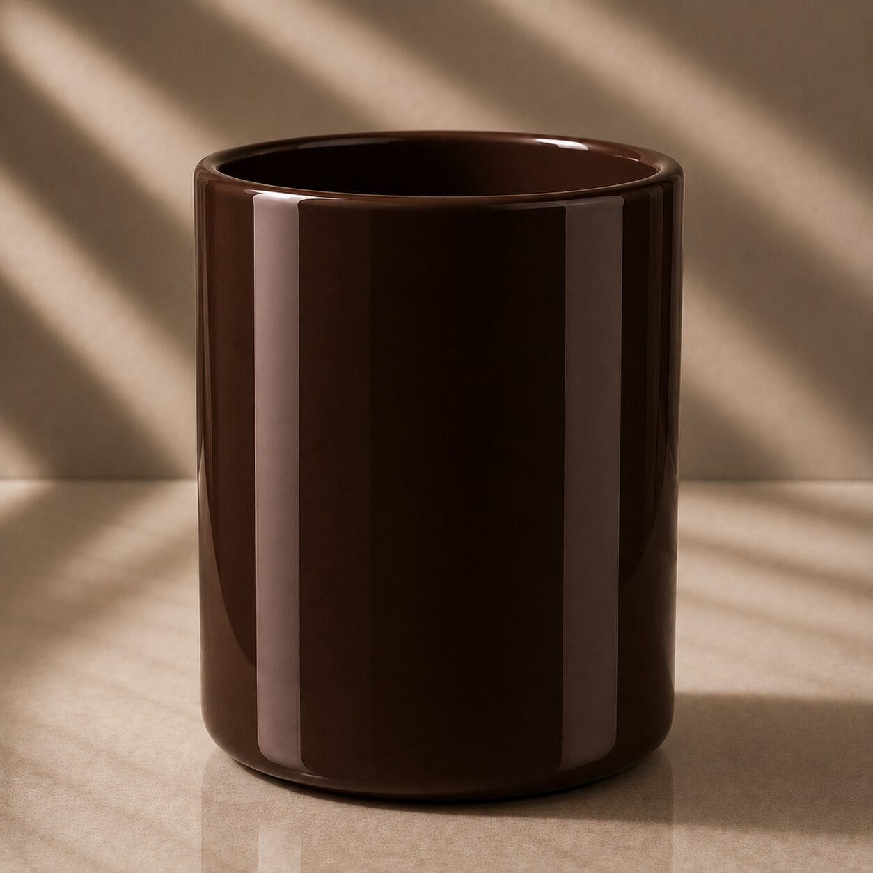 Dark brown glossy cylindrical mug or candle jar with smooth curved sides, displayed on neutral beige surface with diagonal sunlight casting shadows