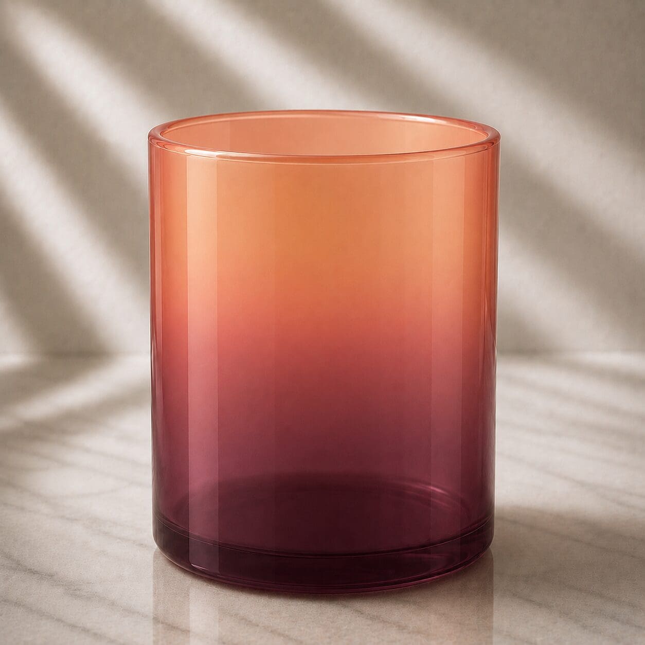 Gradient-colored glass cylinder in coral and burgundy tones on marble surface with natural light casting shadows