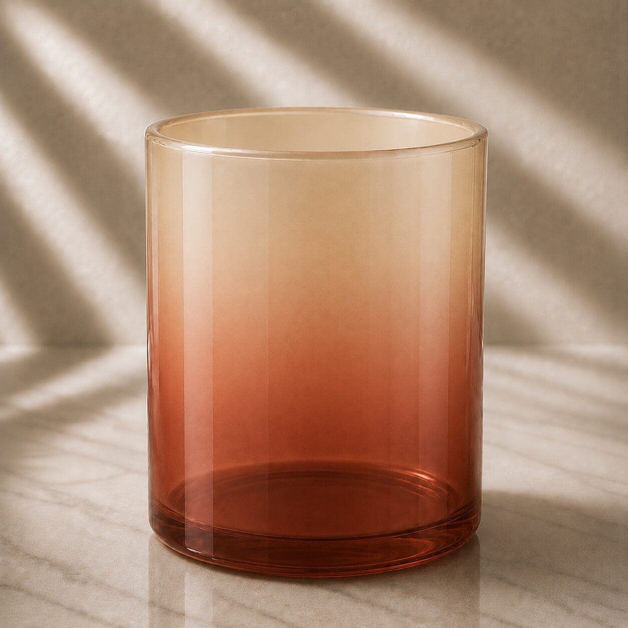 Clear glass cylinder with warm amber-to-red ombre gradient, sitting on marble surface with soft natural lighting and shadows