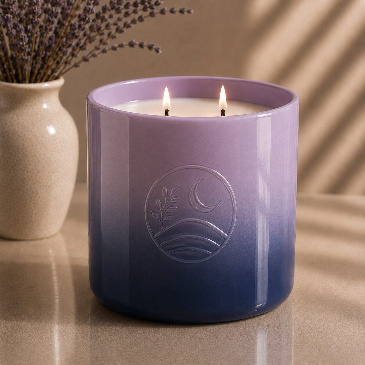 Purple gradient glass candle jar with embossed moon and botanical design, two lit wicks, beside cream ceramic vase with dried lavender