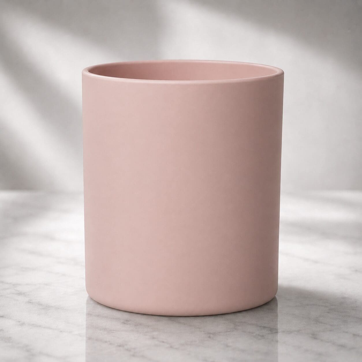 A plain pink ceramic cylinder sits on a marble surface with soft natural light and shadows in the background, suitable for candle filling or decorative glass services