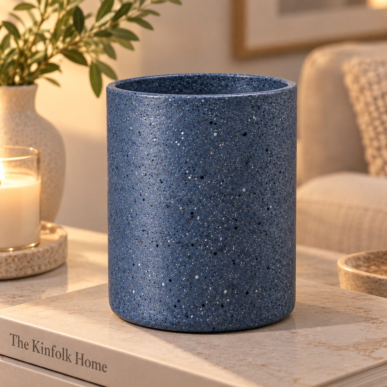Blue speckled ceramic cylinder container on wooden surface surrounded by candles, plants, and home decor items