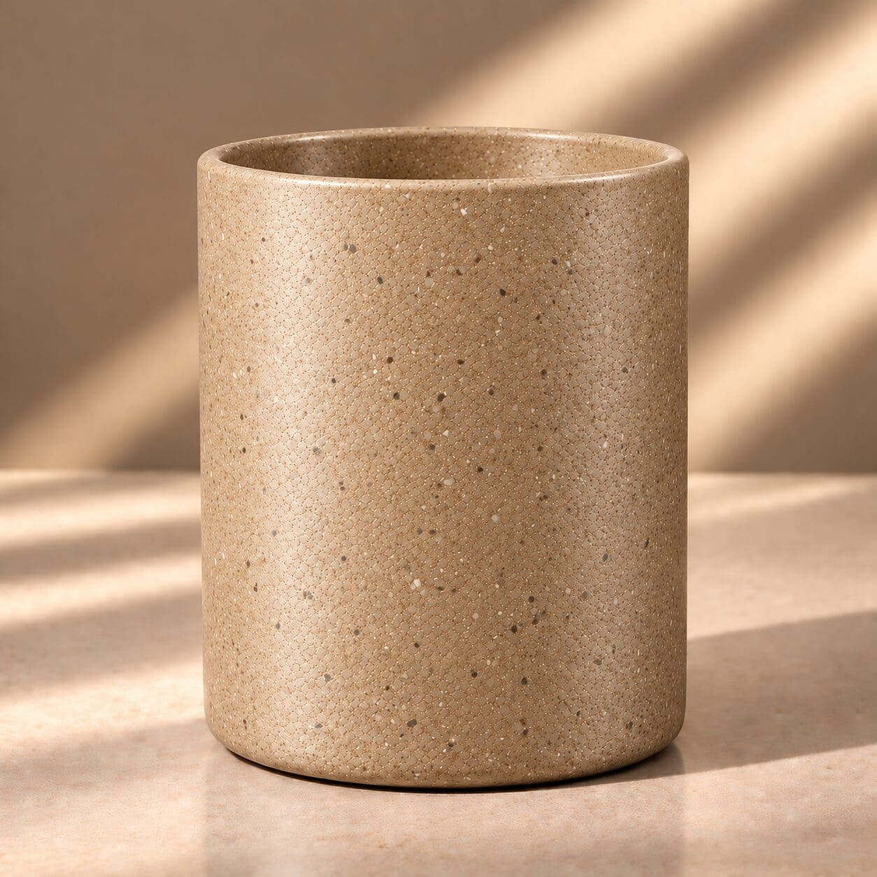 Beige speckled ceramic cylinder cup with textured surface sitting on neutral background with soft shadows