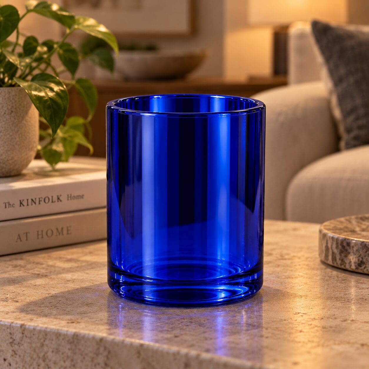 Cobalt blue glass cylinder on wooden surface with books, potted plant, and home decor in soft lighting