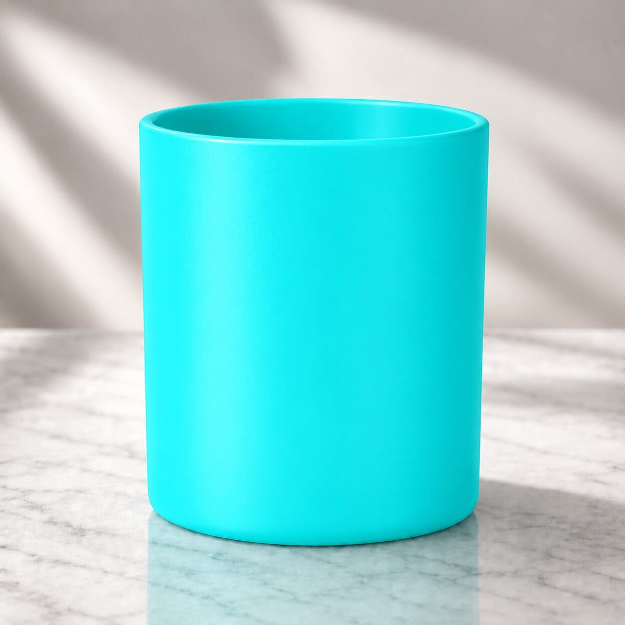 Bright turquoise cylindrical glass jar displayed on white marble surface with natural sunlight casting shadows in background