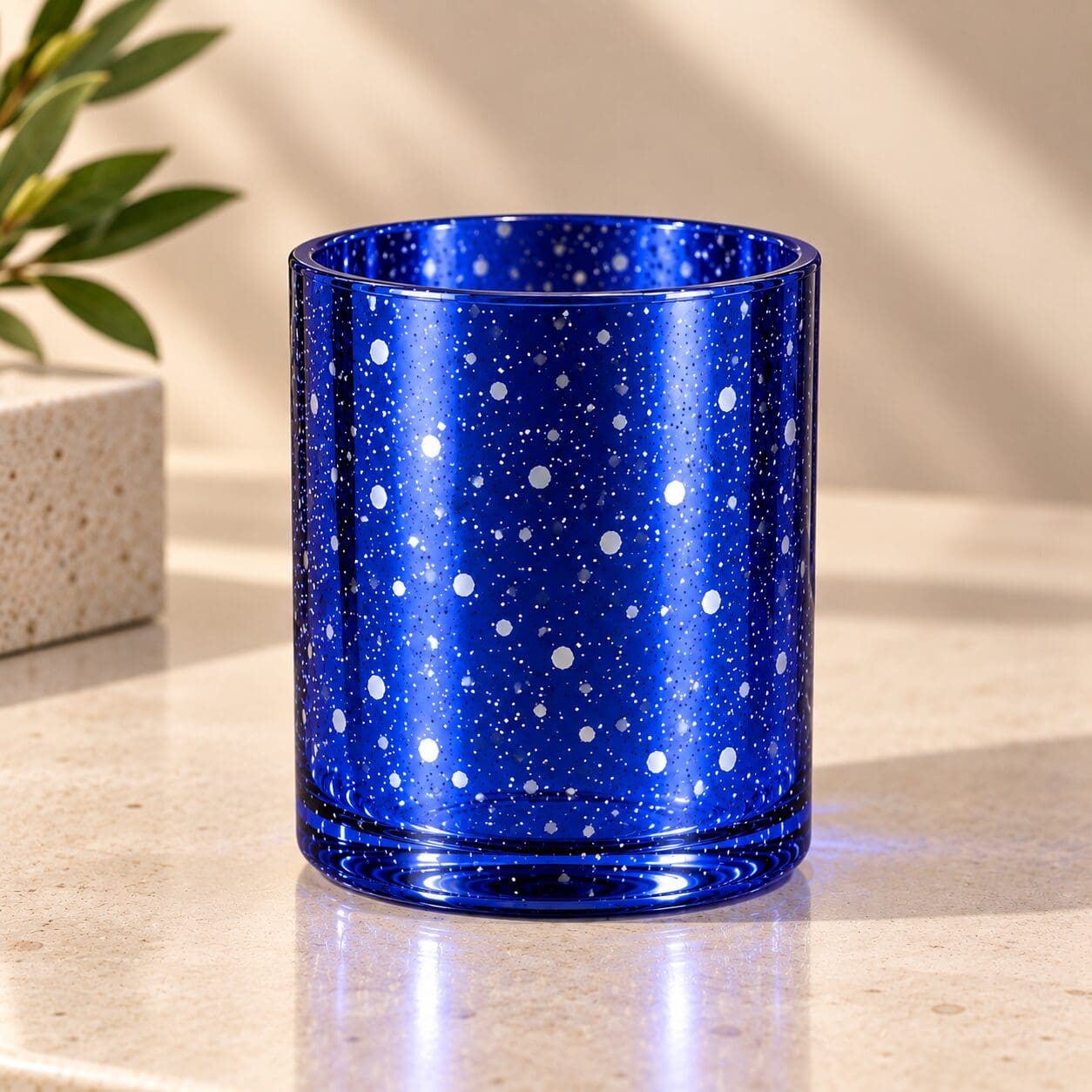 Cobalt blue glass cylinder with white speckled spray coating design, displayed on marble surface next to potted plant