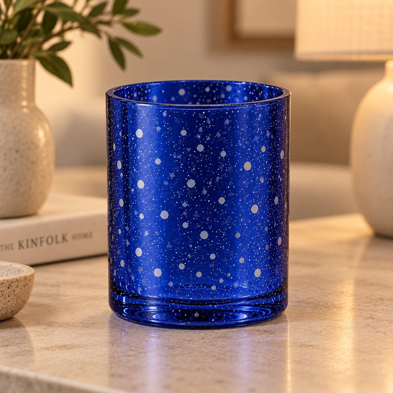 Deep blue speckled glass cylinder with white and gray dots, displayed on marble surface next to potted plant and book, exemplifying decorative glass spray coating and candle jar design