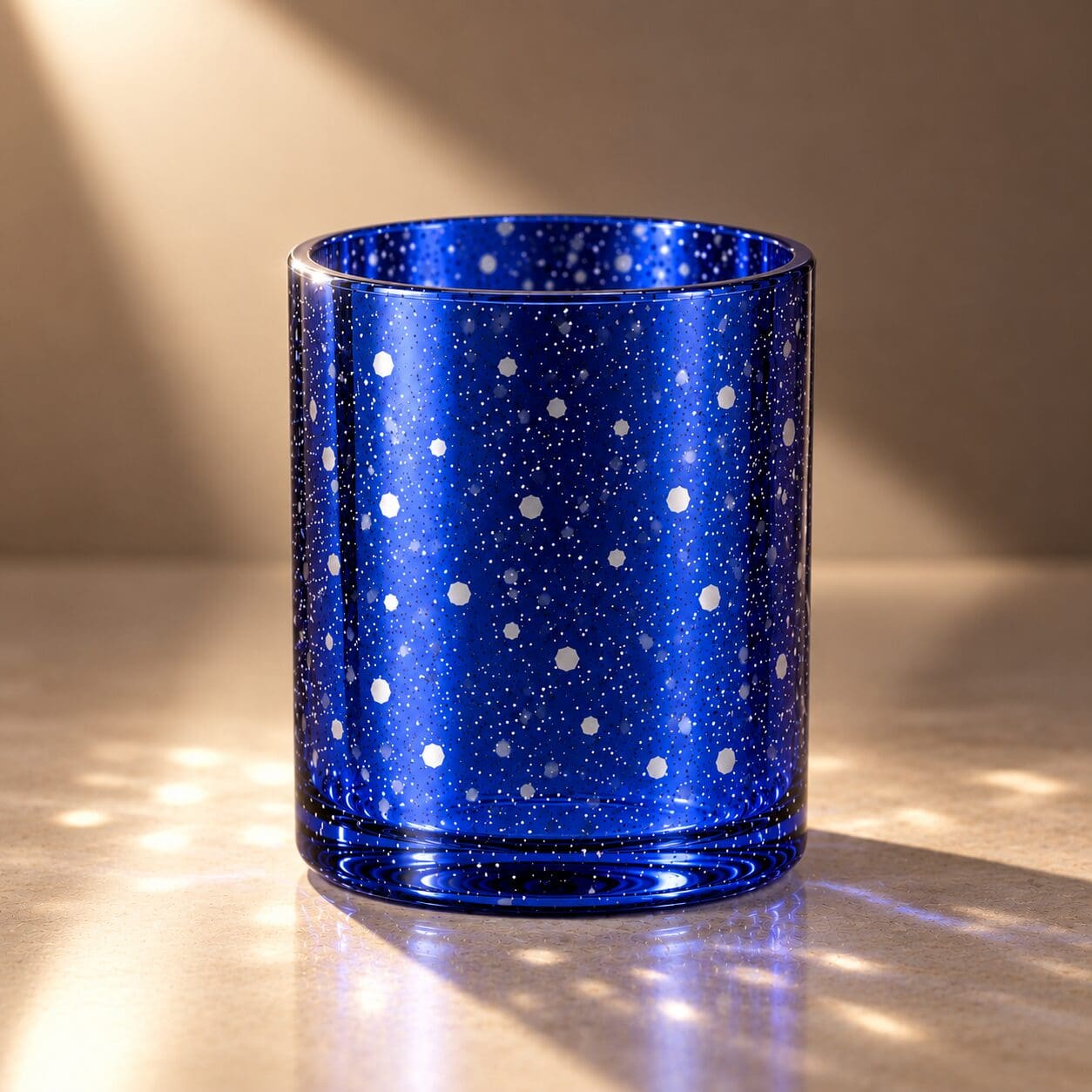 Deep blue glass cylinder with white and silver speckled pattern, illuminated by warm sunlight casting shadows on neutral surface