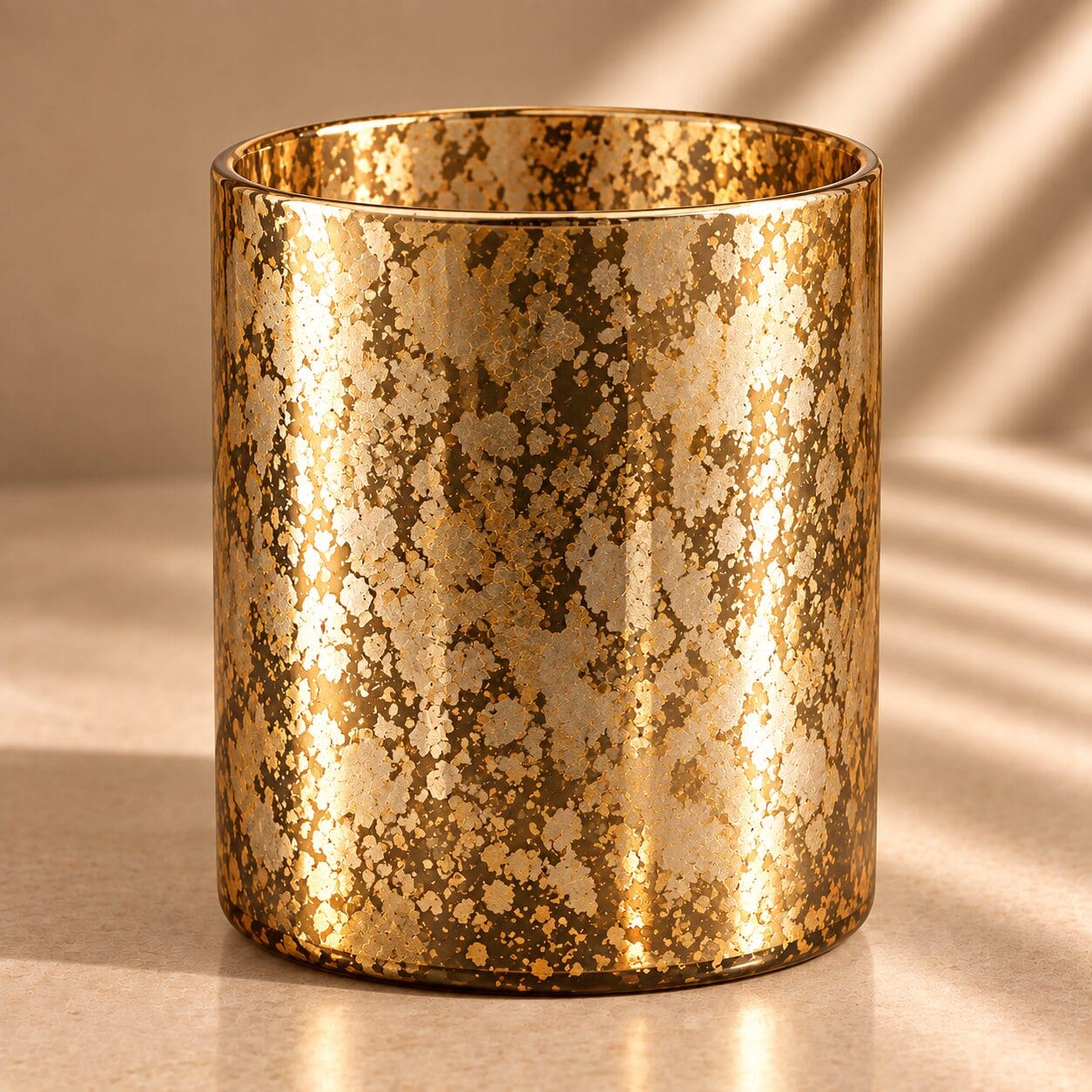 Gold glitter spray coated cylindrical glass jar with speckled metallic finish, casting shadows on beige surface