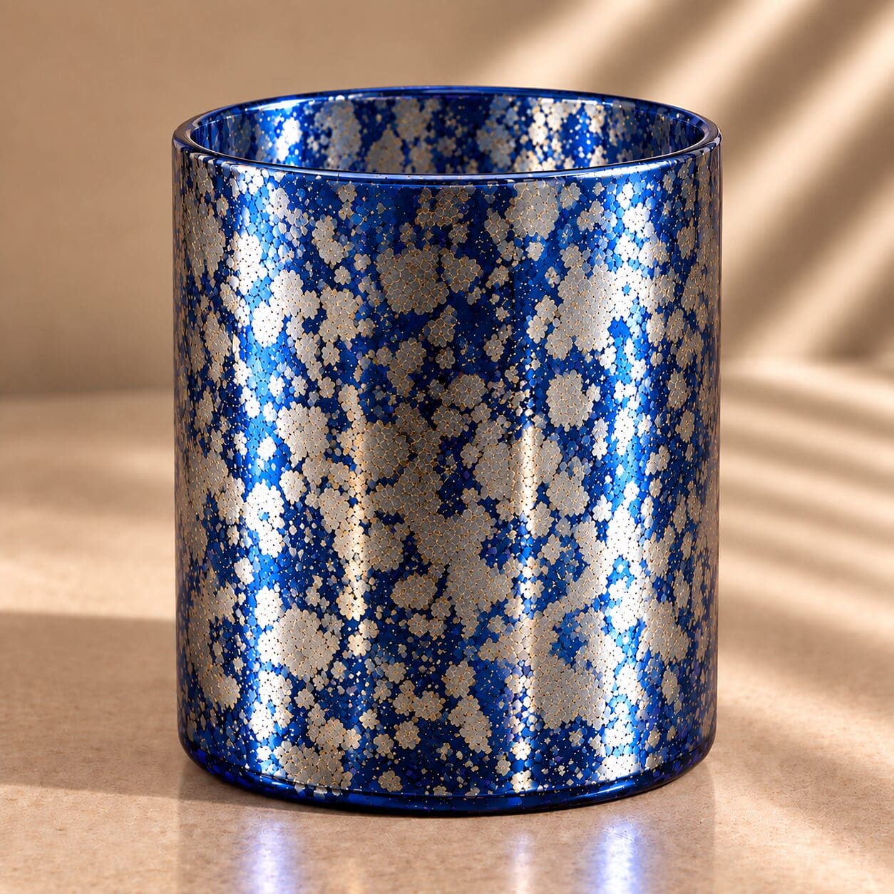 Cylindrical glass jar with spray-coated navy blue and metallic gold speckled finish, catching sunlight on neutral background