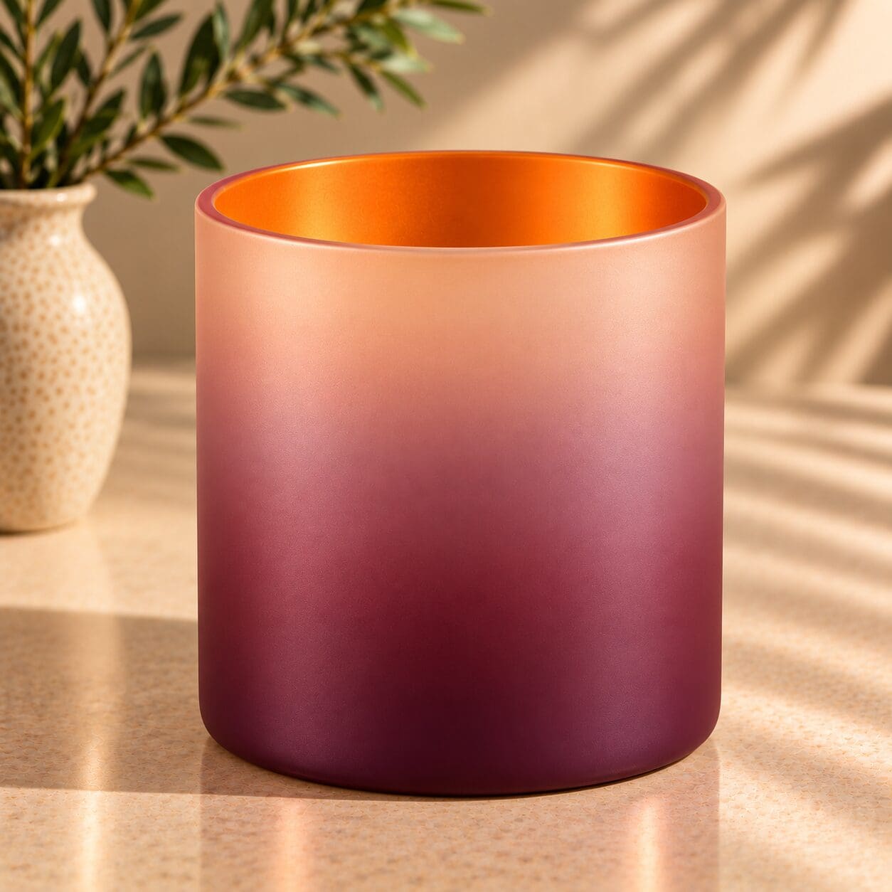 Gradient spray-coated glass cylinder with peachy-orange top fading to deep burgundy, displaying spray coating and glass decorating techniques, positioned on wooden surface next to potted plant