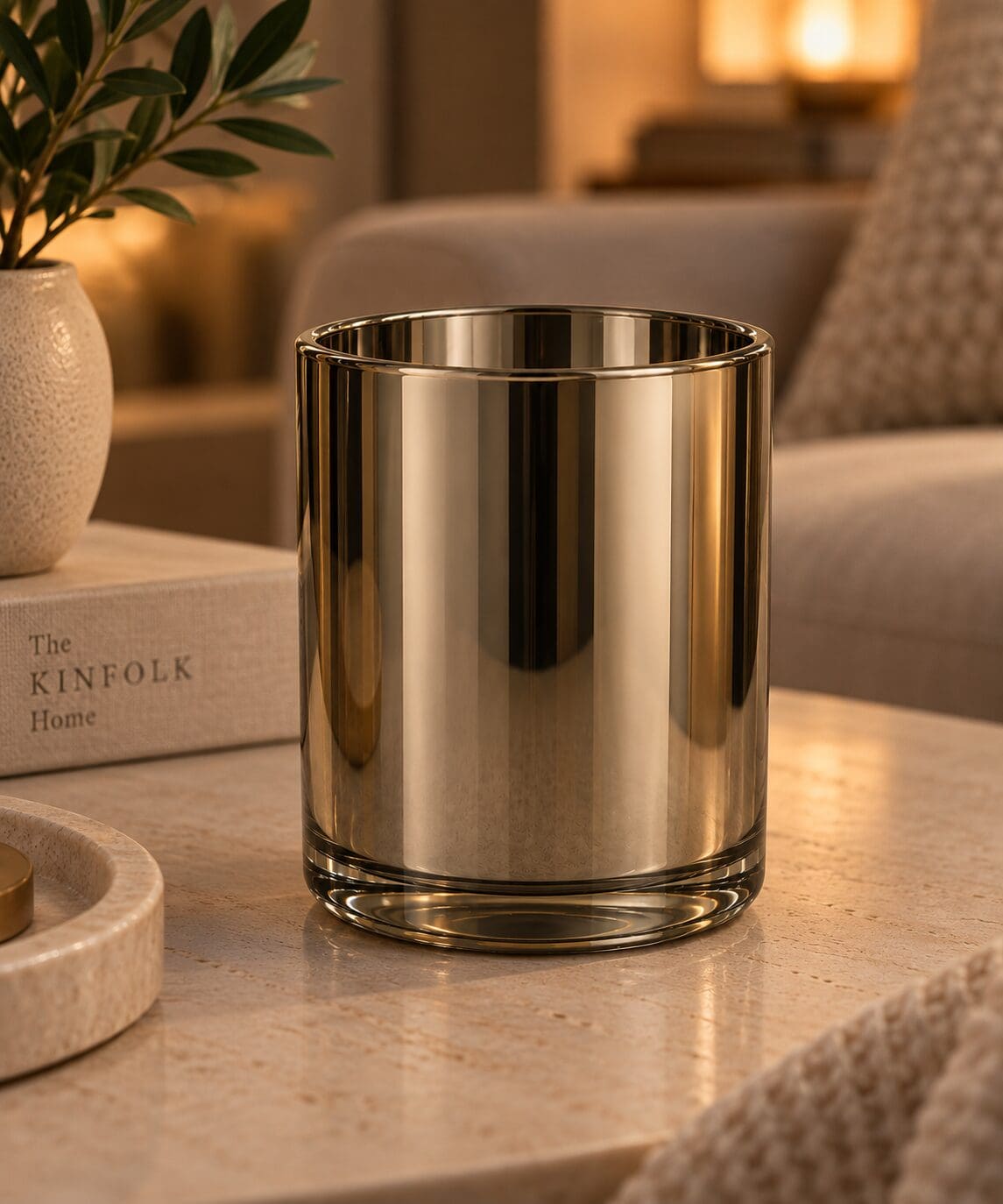 Gold metallic cylindrical glass candle jar on wooden table with plant and home decor in warm-lit living room setting